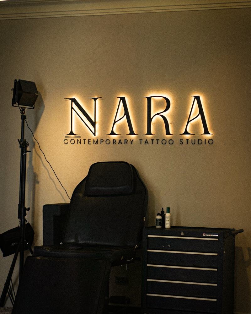 Letrero retroiluminado de NARA Contemporary Tattoo Studio sobre la butaca de trabajo y el carro de material.