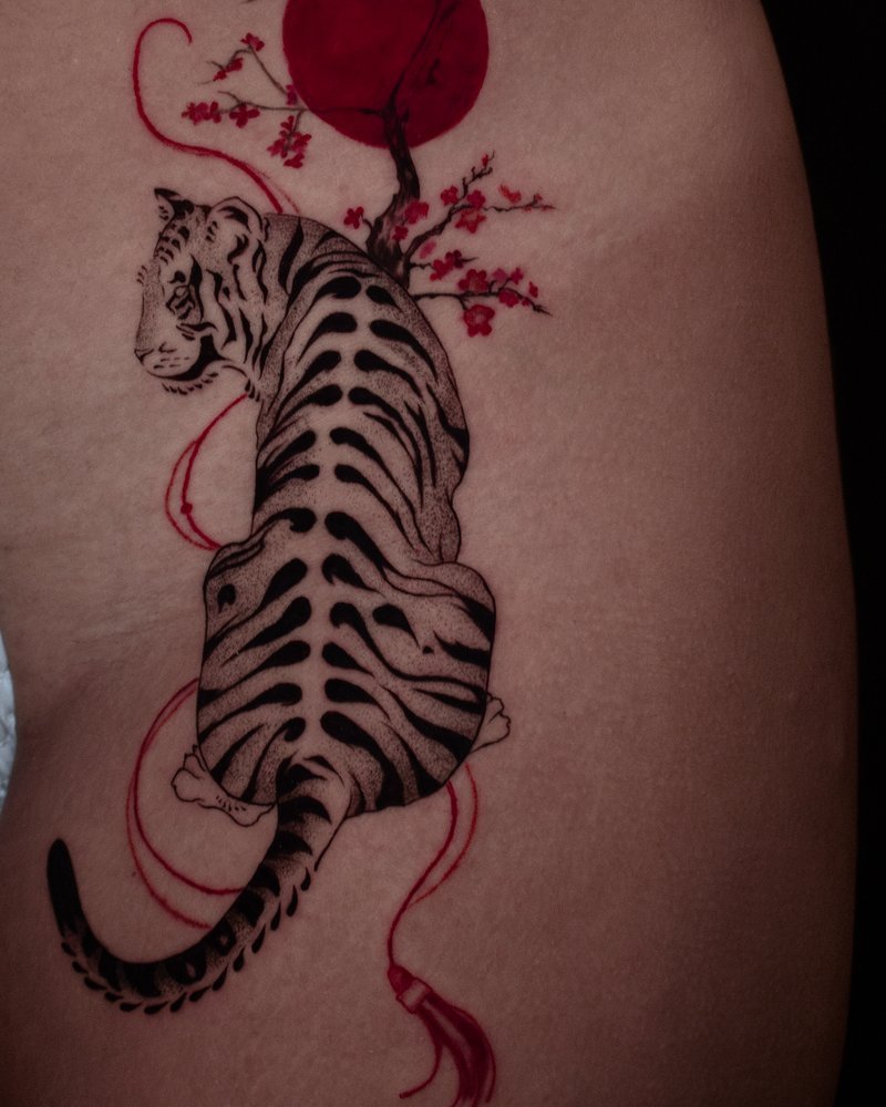 Pieza de Melody Goris: tigre entre ramas de cerezo con un sol en tinta roja, cadera
